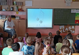 Uczniowie siedzą przed tablicą multimedialną.