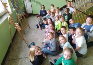 Uczniowie siedzą przed sztalugami ze zdjęciami nauczycieli
